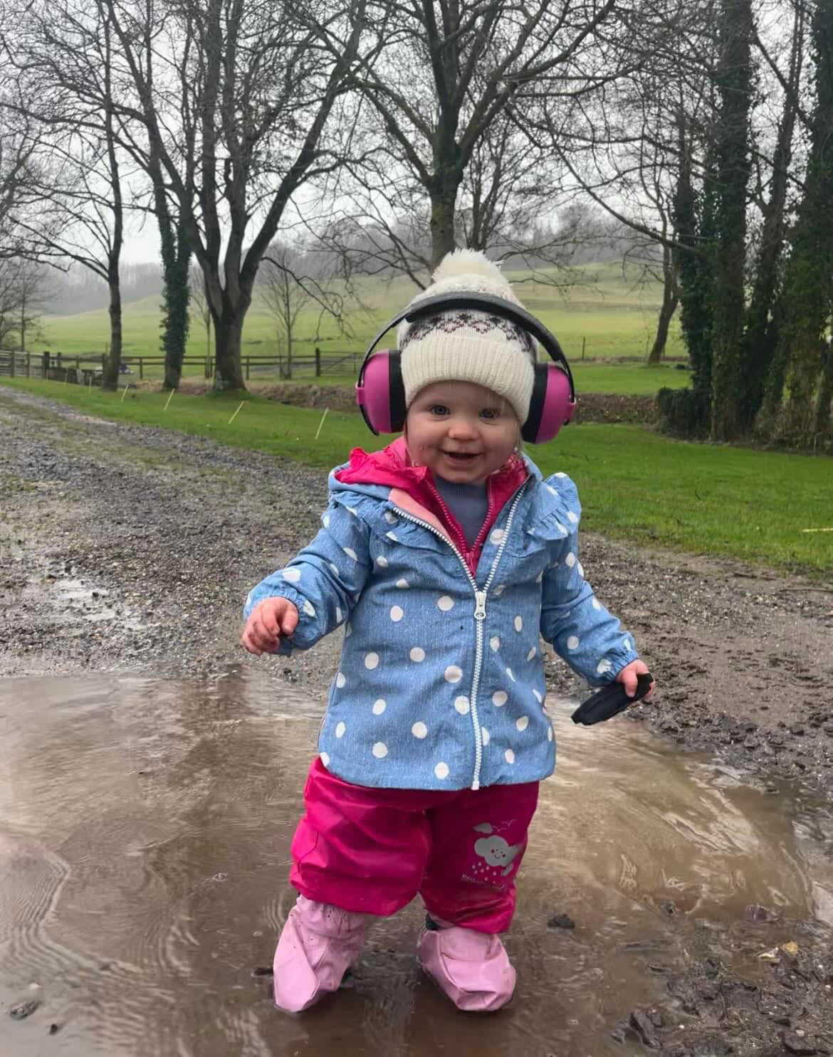 A happy toddler wearing magenta BANZ baby ear muffs and a bobble hat, splashing in a puddle